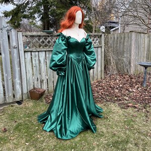 Puede incluir: Un vestido largo de sat&eacute;n verde esmeralda con escote de coraz&oacute;n y mangas abullonadas grandes. El vestido tiene un corpi&ntilde;o ajustado y una falda fluida.