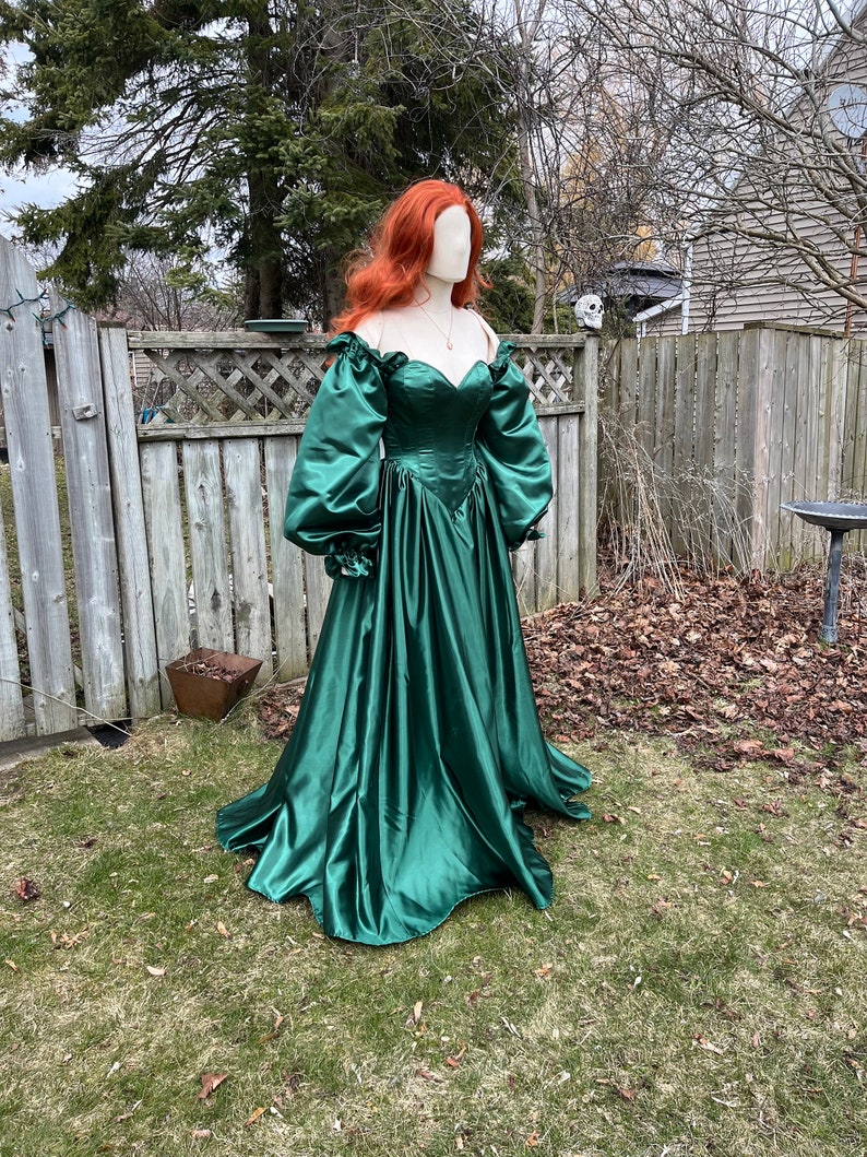 Puede incluir: Un vestido largo de sat&eacute;n verde esmeralda con escote de coraz&oacute;n y mangas abullonadas grandes. El vestido tiene un corpi&ntilde;o ajustado y una falda fluida.