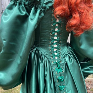 Puede incluir: Un vestido de sat&eacute;n verde con un corpi&ntilde;o ajustado y una falda completa. El corpi&ntilde;o tiene una espalda con cordones con cinta blanca y ojales dorados.