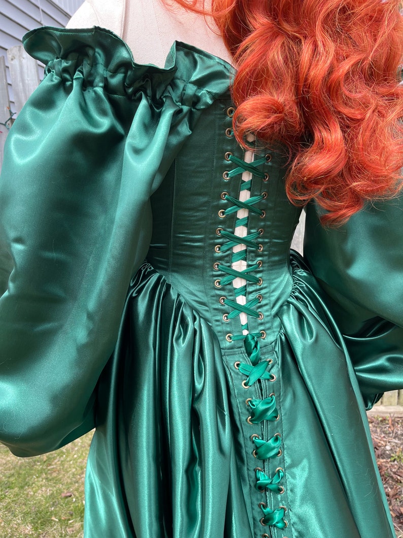 Puede incluir: Un vestido de sat&eacute;n verde con un corpi&ntilde;o ajustado y una falda completa. El corpi&ntilde;o tiene una espalda con cordones con cinta blanca y ojales dorados.