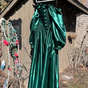 Puede incluir: Un vestido largo de sat&eacute;n verde esmeralda con un corpi&ntilde;o ajustado y una falda completa. El vestido tiene un escote con volantes y mangas largas abullonadas con volantes en los pu&ntilde;os. El corpi&ntilde;o tiene un cierre de encaje en la parte delantera.