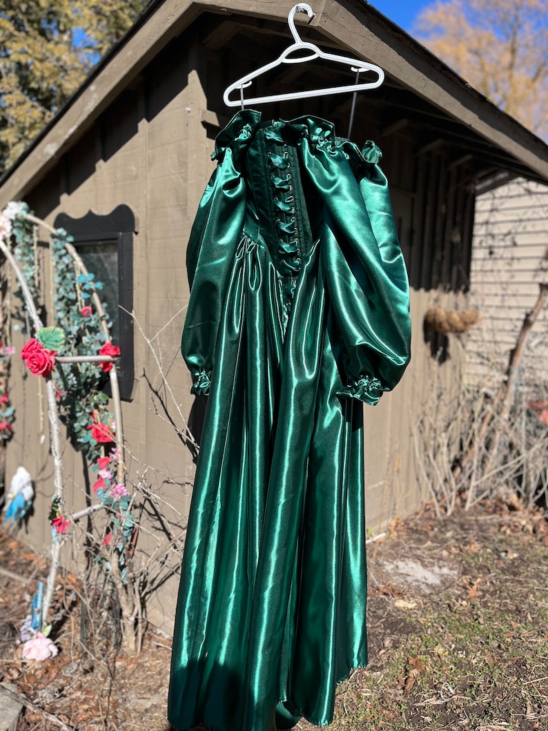 Puede incluir: Un vestido largo de sat&eacute;n verde esmeralda con un corpi&ntilde;o ajustado y una falda completa. El vestido tiene un escote con volantes y mangas largas abullonadas con volantes en los pu&ntilde;os. El corpi&ntilde;o tiene un cierre de encaje en la parte delantera.