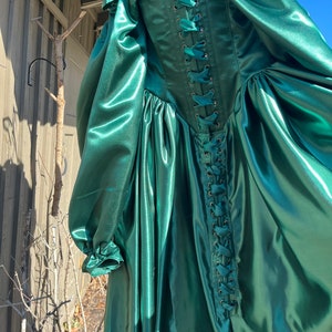 Puede incluir: Un vestido largo de sat&eacute;n verde esmeralda con la espalda con cordones. El vestido tiene un corpi&ntilde;o ajustado y una falda completa.