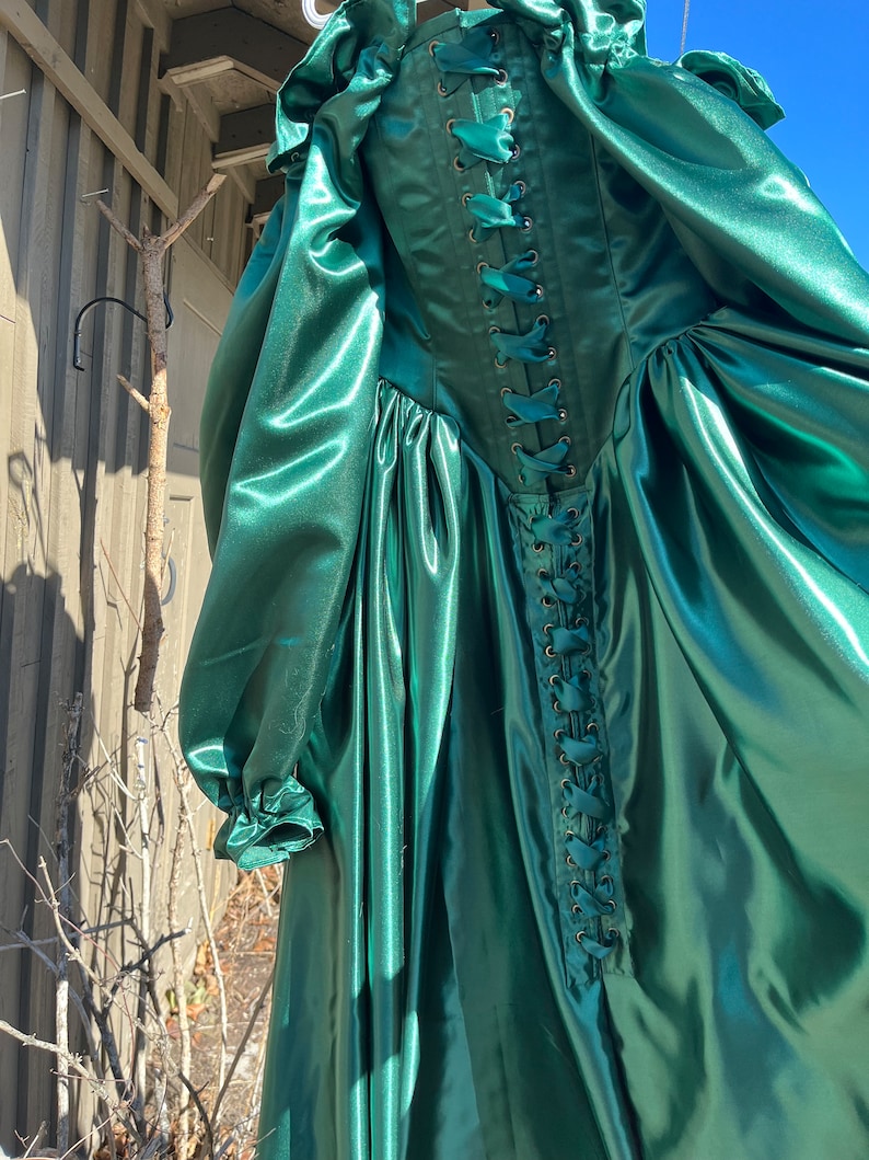 Puede incluir: Un vestido largo de sat&eacute;n verde esmeralda con la espalda con cordones. El vestido tiene un corpi&ntilde;o ajustado y una falda completa.
