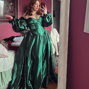 Puede incluir: Una mujer con un vestido verde de sat&eacute;n de hombros descubiertos con mangas largas y abullonadas. El vestido tiene un corpi&ntilde;o ajustado y una falda fluida. La mujer est&aacute; de pie frente a un espejo, y la habitaci&oacute;n est&aacute; decorada en un esquema de color rosa y blanco.