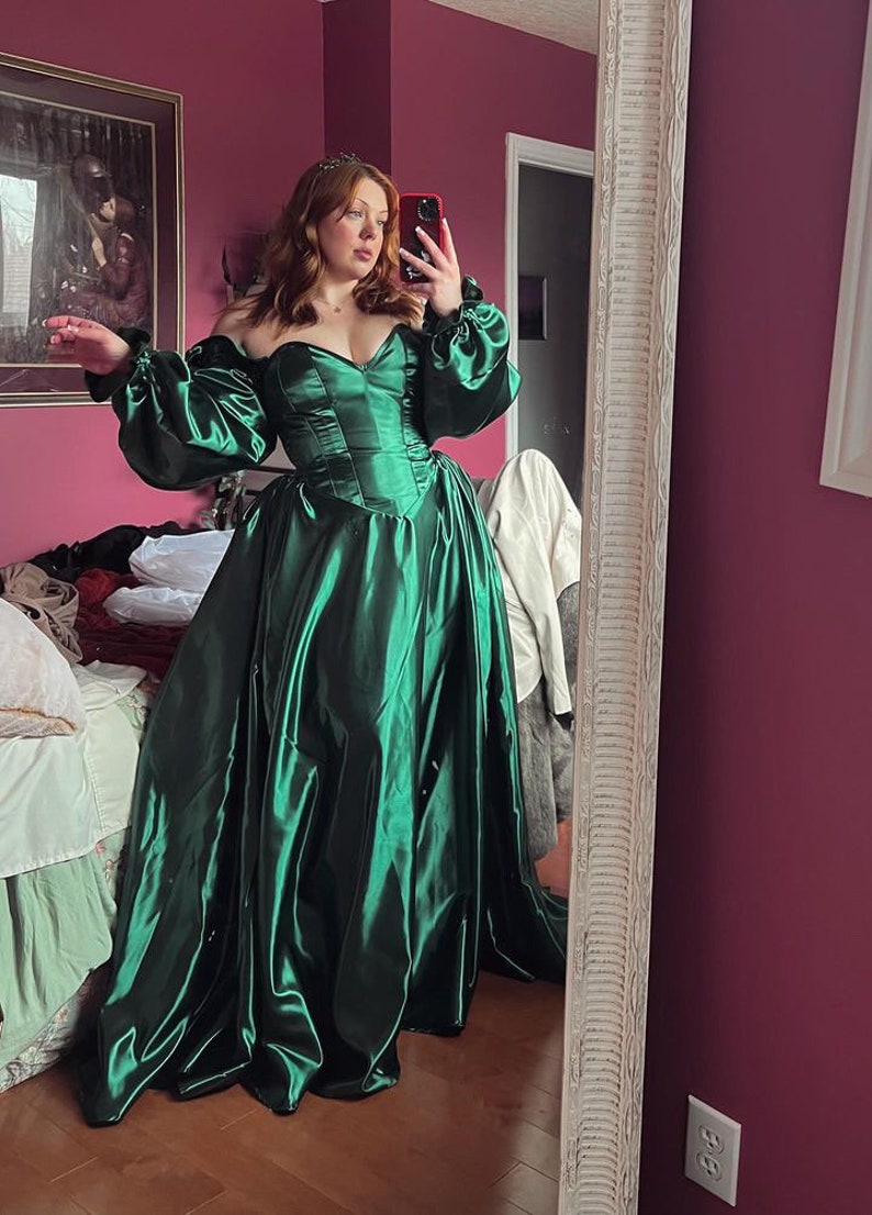 Puede incluir: Una mujer con un vestido verde de sat&eacute;n de hombros descubiertos con mangas largas y abullonadas. El vestido tiene un corpi&ntilde;o ajustado y una falda fluida. La mujer est&aacute; de pie frente a un espejo, y la habitaci&oacute;n est&aacute; decorada en un esquema de color rosa y blanco.