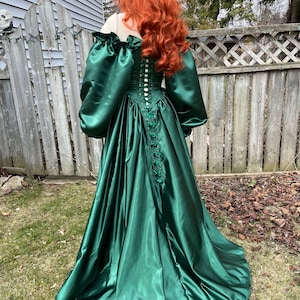 Puede incluir: Un vestido largo de sat&eacute;n verde esmeralda con un corpi&ntilde;o ajustado y una falda completa. El corpi&ntilde;o tiene una espalda con cordones y una fila de botones. El vestido tiene mangas largas y abullonadas.