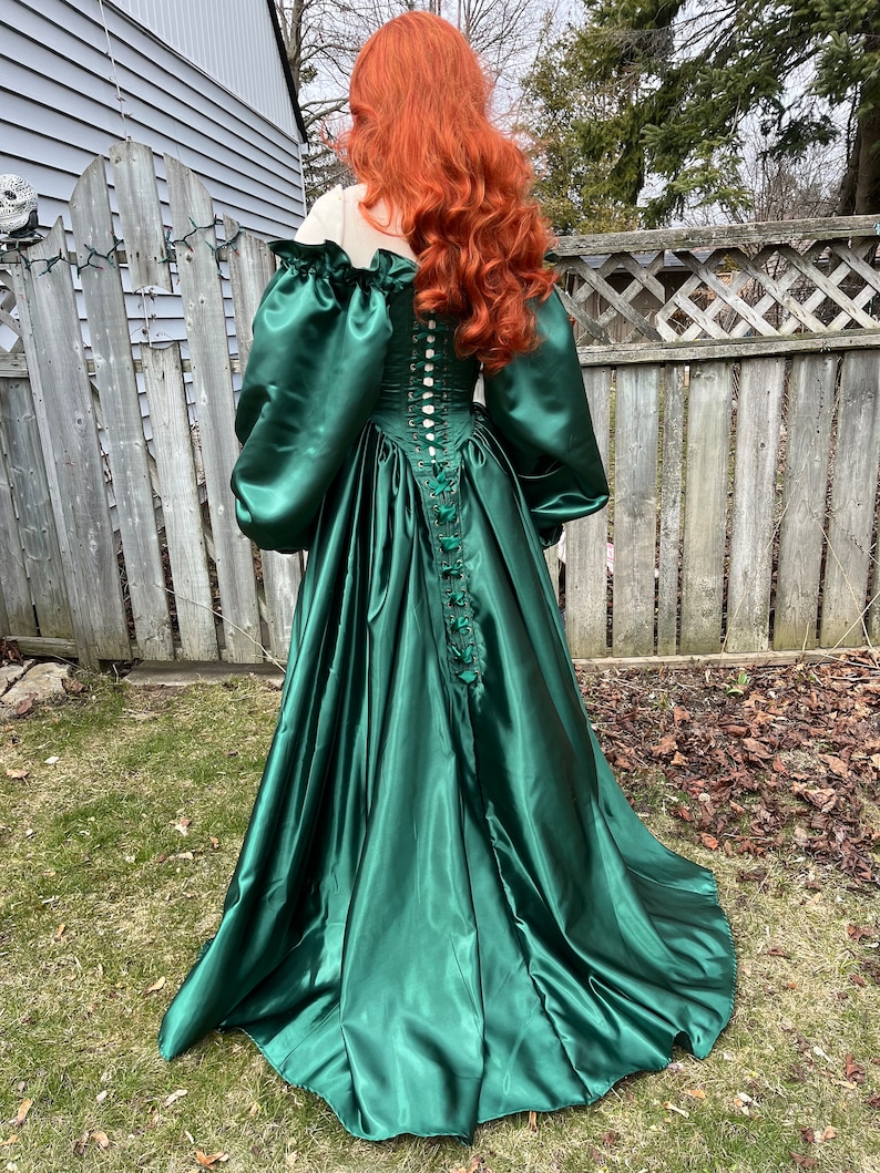 Puede incluir: Un vestido largo de sat&eacute;n verde esmeralda con un corpi&ntilde;o ajustado y una falda completa. El corpi&ntilde;o tiene una espalda con cordones y una fila de botones. El vestido tiene mangas largas y abullonadas.