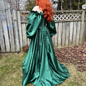 Puede incluir: Un vestido largo de sat&eacute;n verde esmeralda con un corpi&ntilde;o ajustado y una falda completa. El vestido tiene una espalda con cordones y mangas abullonadas.