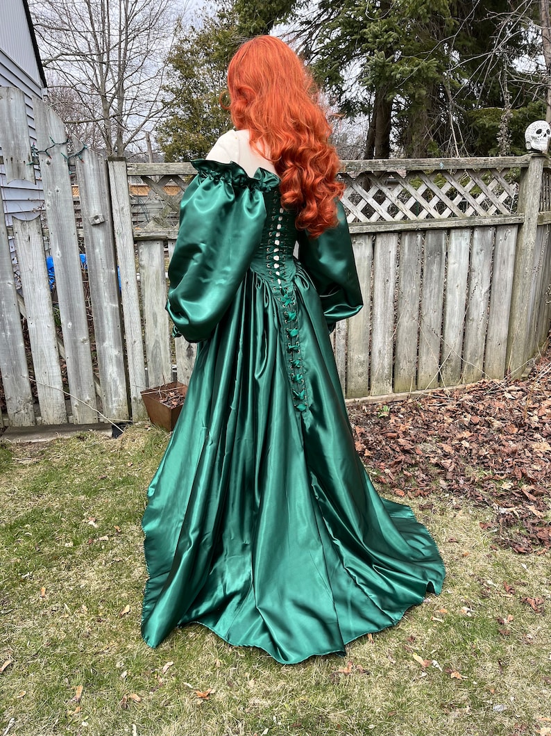 Puede incluir: Un vestido largo de sat&eacute;n verde esmeralda con un corpi&ntilde;o ajustado y una falda completa. El vestido tiene una espalda con cordones y mangas abullonadas.