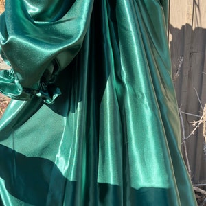 Puede incluir: Una tela de sat&eacute;n verde oscuro con una textura lisa y brillante. La tela est&aacute; drapeada y doblada, creando l&iacute;neas suaves y fluidas.