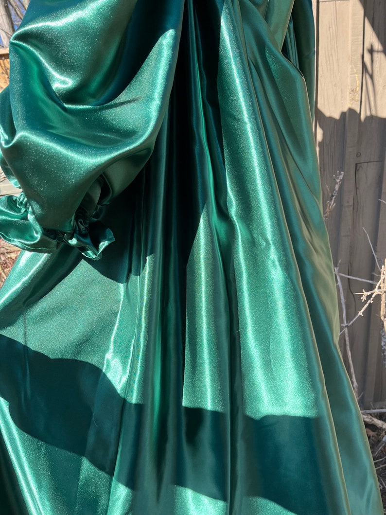 Puede incluir: Una tela de sat&eacute;n verde oscuro con una textura lisa y brillante. La tela est&aacute; drapeada y doblada, creando l&iacute;neas suaves y fluidas.