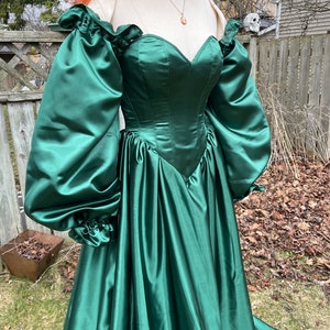 Puede incluir: Un vestido de sat&eacute;n verde con escote de coraz&oacute;n y mangas largas y abullonadas. El vestido tiene un corpi&ntilde;o ajustado y una falda fluida.