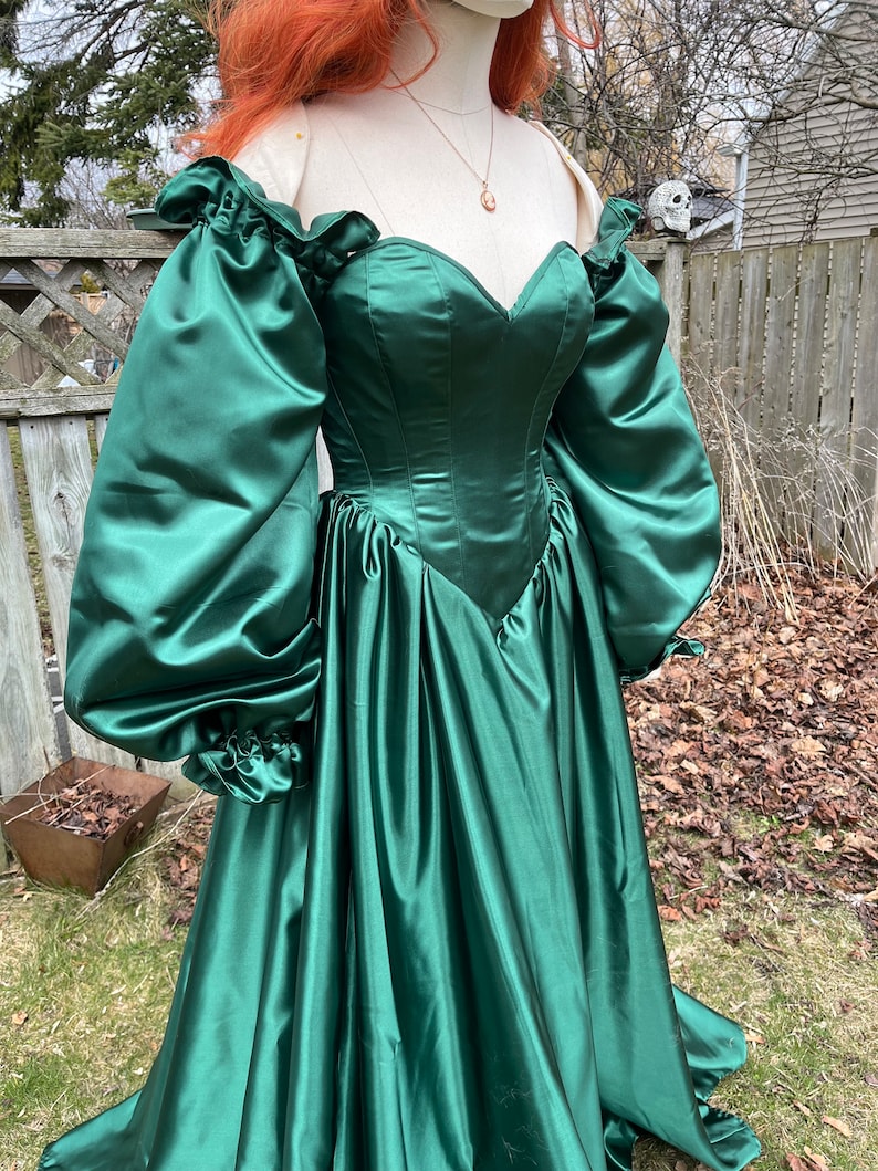 Puede incluir: Un vestido de sat&eacute;n verde con escote de coraz&oacute;n y mangas largas y abullonadas. El vestido tiene un corpi&ntilde;o ajustado y una falda fluida.