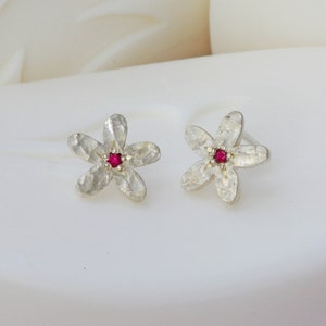 Può includere: Un paio di orecchini a bottone in argento a forma di fiore con una piccola pietra rossa al centro di ogni fiore.