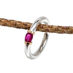 May include: A polished silver ring featuring a smooth, rounded band with a 925 hallmark stamp. The center of the ring is accented by a vibrant, oval-shaped magenta gemstone held securely between two small, gold-toned metal bands.