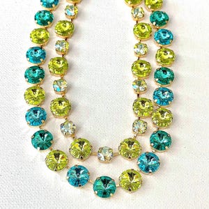 May include: A double-strand necklace featuring round, faceted crystals in alternating shades of lime green and turquoise blue. The crystals are set in gold-toned metal settings and connected by thin gold-toned metal links in a linear arrangement.