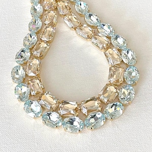 May include: A layered necklace featuring alternating rows of light blue and champagne-colored rectangular and oval-cut crystals. The crystals are set in a gold-toned metal. The necklace is displayed on a white surface.