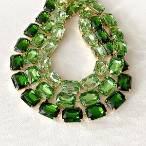 May include: A teardrop-shaped statement necklace featuring multiple rows of faceted rectangular crystals in varying shades of green, ranging from light lime to deep forest green, set in a gold-toned metal frame with a secure link construction.
