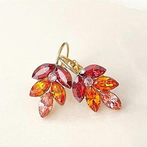 May include: A pair of gold-tone earrings featuring a cluster of marquise-cut crystals in shades of deep red, bright orange, and light pink. Each earring is designed with a central clear stone surrounded by leaf-shaped crystals and a lever-back closure.