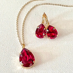 May include: A gold-toned necklace with a teardrop-shaped, ruby-red pendant and matching earrings. The earrings and pendant feature a vibrant, faceted gemstone set in a gold frame. The jewelry is displayed on a white, textured surface.