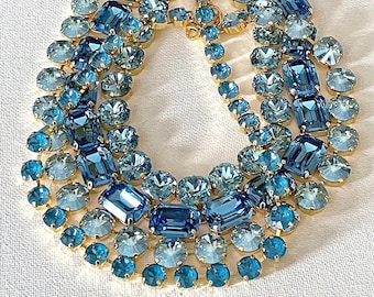 May include: A multi-strand statement necklace featuring an array of faceted blue and light blue crystals in various shapes, including round and rectangular cuts, set in gold-toned metal settings that create a dense, layered, and sparkling appearance.