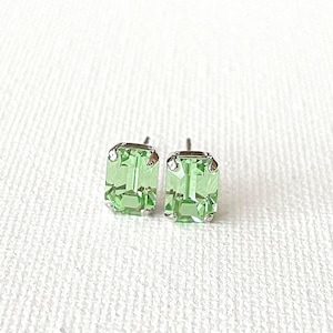 May include: A pair of light green emerald-cut stud earrings. The earrings have a silver-colored setting and a faceted, rectangular green gemstone. The earrings are displayed on a white, textured surface.