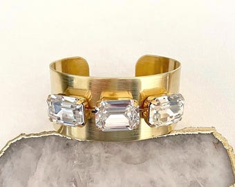 May include: A gold-toned cuff bracelet featuring three large, rectangular, clear crystal stones. The bracelet has a wide band and an open design, showcasing the sparkling crystals.