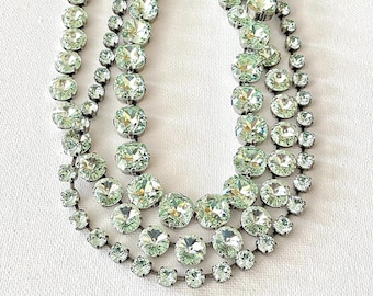 Può includere: Una collana a tre fili con cristalli rotondi verde chiaro. I cristalli sono incastonati in metallo argentato. La collana è esposta su una superficie bianca. Un gioiello importante.