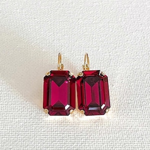 May include: A pair of ruby red rectangular drop earrings with gold-colored metal accents. The earrings have a faceted design, catching the light. The lever-back closures are also gold-colored. These earrings are a statement piece.