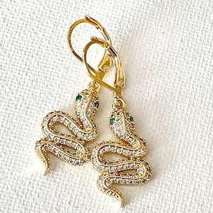 May include: Gold-toned snake earrings with clear and green crystal accents. The earrings feature a lever-back closure and are designed in the shape of a coiled snake. The snake's body is covered in small, clear crystals, and the eyes are green.