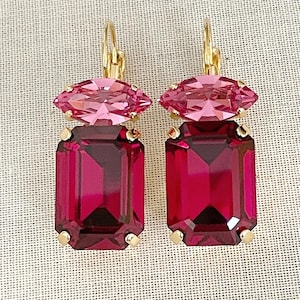 May include: A pair of gold-toned earrings featuring a light pink marquise-cut crystal above a large, rectangular, deep magenta crystal. The earrings have a lever-back closure and are set against a neutral background.