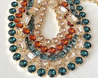 May include: A multi-strand statement necklace featuring gold-tone metal settings holding faceted crystals in shades of deep teal, burnt orange, and champagne. The design includes round and rectangular stones arranged in a layered, cascading pattern.