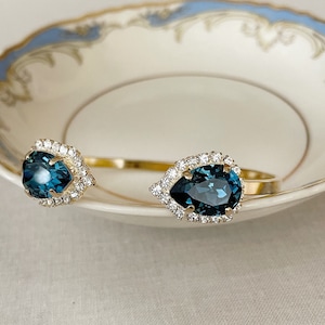 May include: A gold bangle bracelet with two teardrop-shaped blue gemstones surrounded by clear rhinestones.