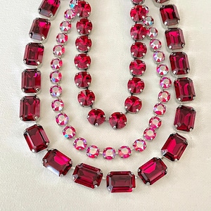 May include: Three strands of ruby-red gemstone necklaces. The top and bottom strands feature large, rectangular stones, while the middle strand has smaller, round stones. The stones are set in silver-toned metal.
