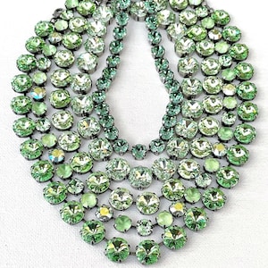 May include: A multi-strand statement necklace featuring numerous round, faceted light green crystals set in dark metal settings. The design consists of several cascading rows of varying sizes of circular stones arranged in a dense, layered pattern.