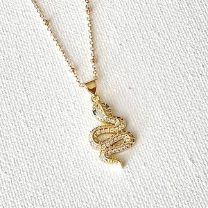 May include: Gold-toned snake pendant necklace. The pendant is encrusted with clear crystals and features a green crystal eye. The chain is delicate with small beaded accents. The necklace is displayed on a white surface.