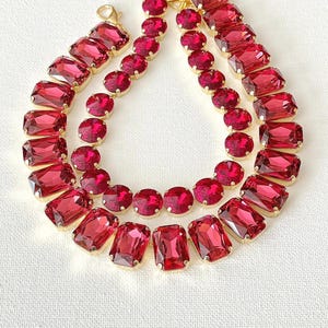 May include: A double-strand necklace featuring faceted, rectangular and circular deep red crystals set in gold-toned metal settings. The necklace includes a lobster claw clasp and is arranged in a layered, circular formation on a textured surface.
