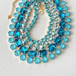 May include: A multi-strand statement necklace featuring a variety of blue and turquoise crystals and gemstones. The necklace has a gold-tone chain and clasp. The gemstones are in various shapes, including round, rectangular, and faceted.