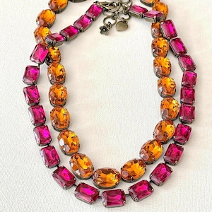 May include: A two-strand necklace featuring alternating oval and rectangular gemstones in shades of orange and magenta. The necklace has a bronze-colored chain and clasp, with small charms. Jewelry.