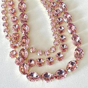 May include: A close-up of three strands of pink crystal necklaces. The crystals are various shapes and sizes, set in gold-colored metal. The necklaces are displayed on a white surface, creating a contrast with the pink crystals.