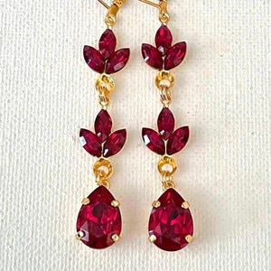 May include: A pair of gold-toned drop earrings with ruby-red gemstones. Each earring features a teardrop-shaped gem at the bottom, a cluster of three leaf-shaped gems, and a smaller cluster at the top. The earrings are displayed against a white background.