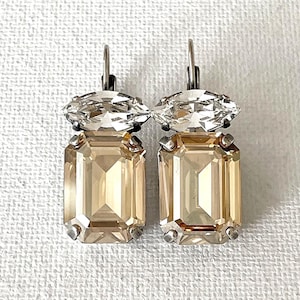 May include: A pair of drop earrings featuring a clear marquise-cut crystal positioned above a larger, rectangular emerald-cut champagne-colored crystal. Each stone is set in a silver-toned metal prong setting with a lever-back closure mechanism.