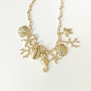 May include: A gold-tone chain necklace featuring multiple dangling charms including a sea turtle, a seahorse, starfish, seashells, and coral branches. Several of the charms are accented with small, clear, round inset stones.