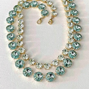 May include: A double-strand necklace featuring round, faceted light blue-green crystals set in gold-toned metal settings. The necklace includes two lobster claw clasps at the ends, with the crystals arranged in a continuous, linked chain pattern.