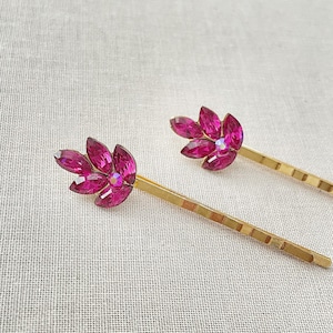 May include: Two gold hair clips with pink jeweled flower designs. Each clip has a row of small gold squares along the length of the clip.