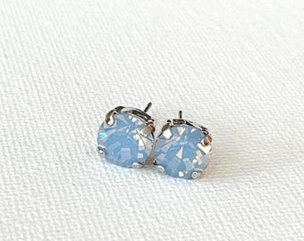 May include: A pair of light blue stud earrings with a faceted, round gemstone design. The earrings have a silver-colored metal setting and are displayed on a white textured surface. The earrings are a jewelry item.