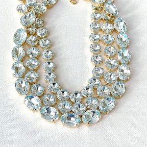 May include: A three-strand necklace featuring clear, oval and round-cut crystals set in gold-tone metal. The crystals are arranged in a graduated design, with larger stones at the bottom and smaller ones towards the top. The necklace is displayed on a white surface.