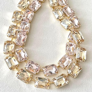 May include: A necklace featuring two rows of rectangular, light champagne-colored faceted crystals set in gold-toned metal frames. The crystals are arranged in a teardrop-shaped pattern, with each stone linked to the next by thin gold-toned metal bars.