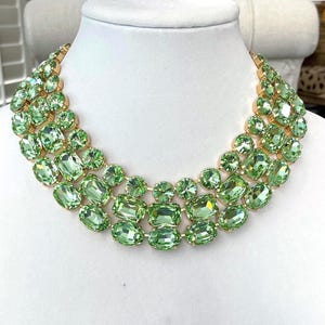 May include: A statement necklace featuring three rows of light green, faceted, oval and rectangular crystals set in a gold-tone metal. The necklace is displayed on a white mannequin bust.